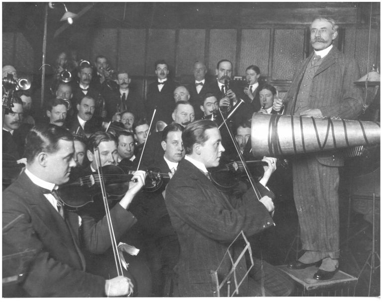 Black and white image featuring composer Edward Elgar surrounded by an orchestra recording music for a gramophone in the early twentieth century