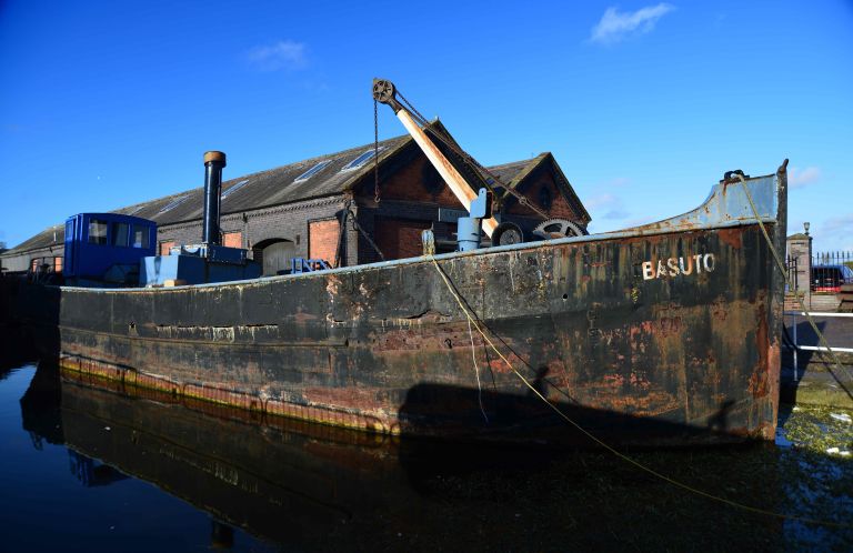 An old, weathered black Clyde puffer named Basuto with a crane on deck, docked in a canal in front of a historic brick warehouse under a clear blue sky.