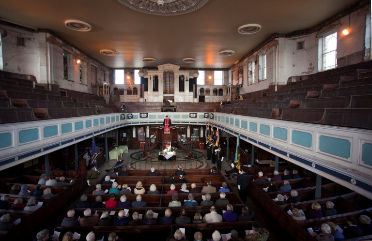 View inside Bethesda Methodist Chapel with worshippers seated in wooden pews beneath a curved balcony, flags near the altar, and warm light.