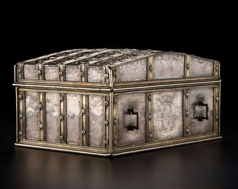 An ornate, rectangular silver chest with a domed lid, featuring intricate floral engravings, metal straps, side handles, and a central coat of arms.