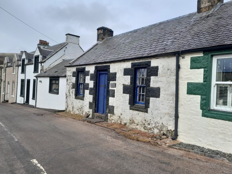 Whitewashed stone cottages line a quiet village street, their slate roofs, blue doors, and dark window trim creating a quaint, historic feel beneath an overcast sky.