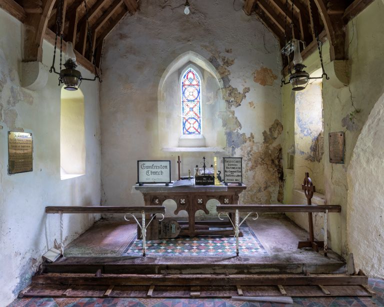 The interior of a rustic church featuring a wooden altar, a stained glass window, worn plaster walls, and a patterned tile floor with wooden pews.