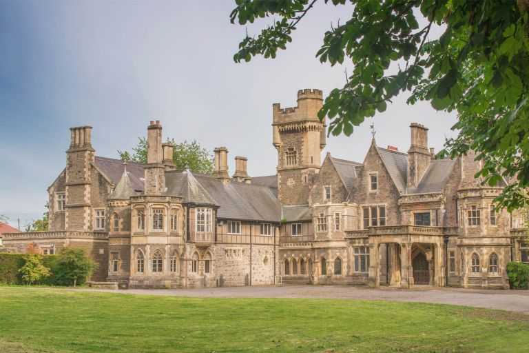 Insole Court Grade II* listed building shown as a large, historic stone manor house with a central tower, multiple chimneys, and arched windows, set behind a green lawn under a pale blue sky.