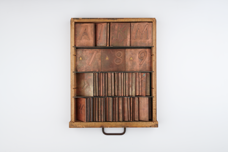 A wooden printer's drawer containing rows of copper letterpress plates, featuring letters, numbers, and symbols organised in four horizontal shelves.