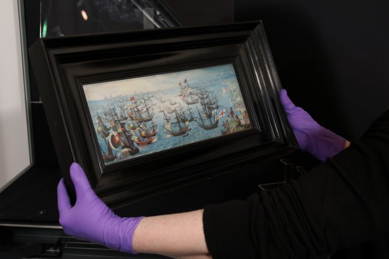 A person wearing purple rubber gloves holds a framed painting of a naval battle featuring several sailing ships on a blue sea against a dark background.