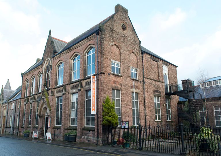 Historic red-brick building with tall arched windows, a peaked roof, and iron railings, photographed on a quiet street beneath a pale sky.