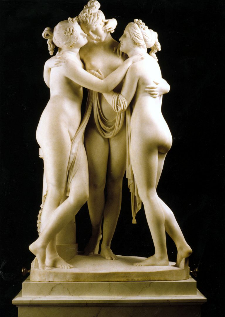 A white marble sculpture of three nude women standing in a circle, embracing each other with draped fabric, set against a solid black background.
