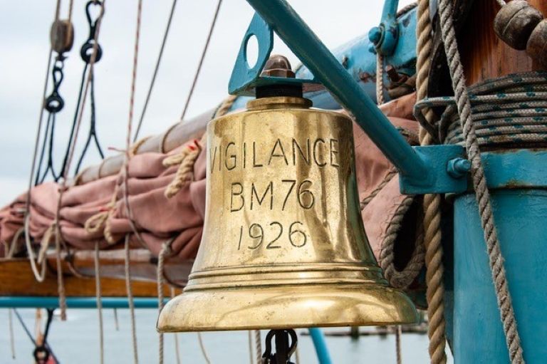Close up of ship's bell on board Brixham heritage trawler Vigilance with ropes, sails and rigging seen in the background. The bell is gold in colour with the words Vigilance, BM76, 1926 engraved on it.
