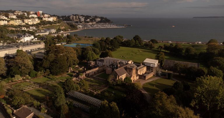 An aerial view of a historic stone abbey building surrounded by gardens and trees, with a coastal town, marina, and the ocean visible in the background.