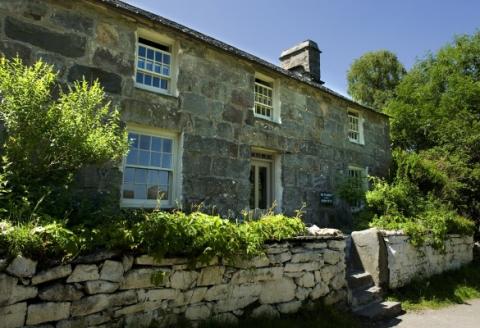 View of a stone built house with sash windows, a chimney and a door. Steps lead up from the road and there are trees and bushes in the surrounding area. 