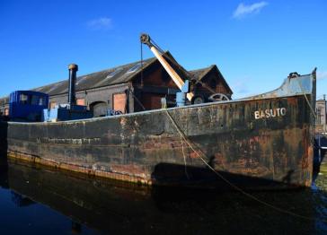 An old, weathered black Clyde puffer named Basuto with a crane on deck, docked in a canal in front of a historic brick warehouse under a clear blue sky.