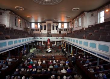View inside Bethesda Methodist Chapel with worshippers seated in wooden pews beneath a curved balcony, flags near the altar, and warm light.