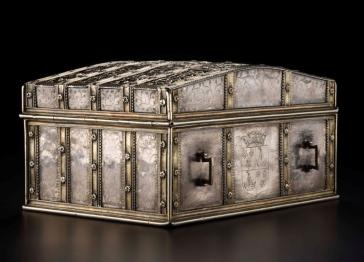 An ornate, rectangular silver chest with a domed lid, featuring intricate floral engravings, metal straps, side handles, and a central coat of arms.