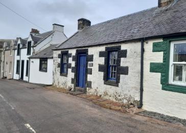 Whitewashed stone cottages line a quiet village street, their slate roofs, blue doors, and dark window trim creating a quaint, historic feel beneath an overcast sky.