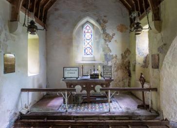 The interior of a rustic church featuring a wooden altar, a stained glass window, worn plaster walls, and a patterned tile floor with wooden pews.