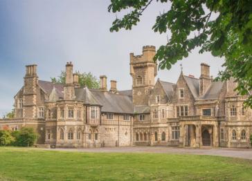 Insole Court Grade II* listed building shown as a large, historic stone manor house with a central tower, multiple chimneys, and arched windows, set behind a green lawn under a pale blue sky.