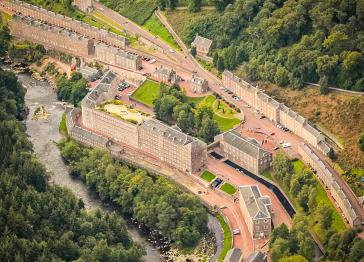 An aerial view of New Lanark, showing historic stone mill buildings and housing rows nestled in a lush, forested valley beside a flowing river.