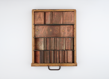 A wooden printer's drawer containing rows of copper letterpress plates, featuring letters, numbers, and symbols organised in four horizontal shelves.