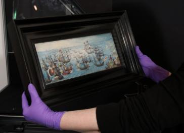A person wearing purple rubber gloves holds a framed painting of a naval battle featuring several sailing ships on a blue sea against a dark background.