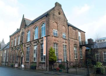Historic red-brick building with tall arched windows, a peaked roof, and iron railings, photographed on a quiet street beneath a pale sky.