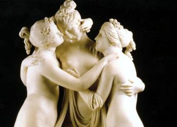 A white marble sculpture of three nude women standing in a circle, embracing each other with draped fabric, set against a solid black background.