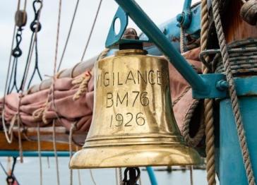Close up of ship's bell on board Brixham heritage trawler Vigilance with ropes, sails and rigging seen in the background. The bell is gold in colour with the words Vigilance, BM76, 1926 engraved on it.