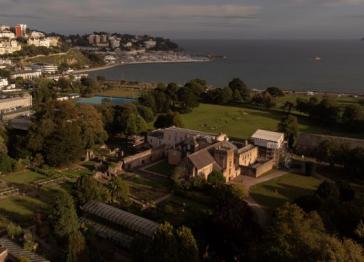 An aerial view of a historic stone abbey building surrounded by gardens and trees, with a coastal town, marina, and the ocean visible in the background.